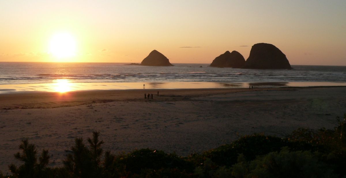 A yellow orange sunset over the ocean with a sandy beach in the foreground and the silhouette of a large rock formation out in the water.