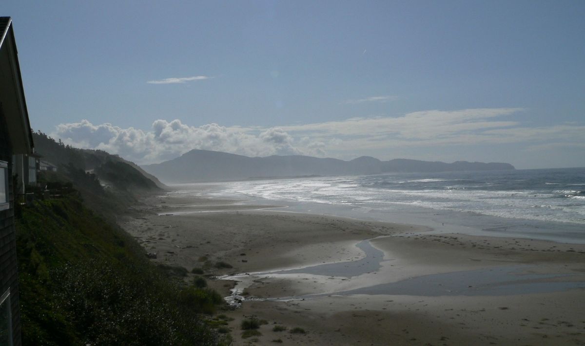 A view down the beach along the coast in Oregon with bright morning light, green hills along the edge of the beach, water pooling on the sand, white crests along the water's edge.