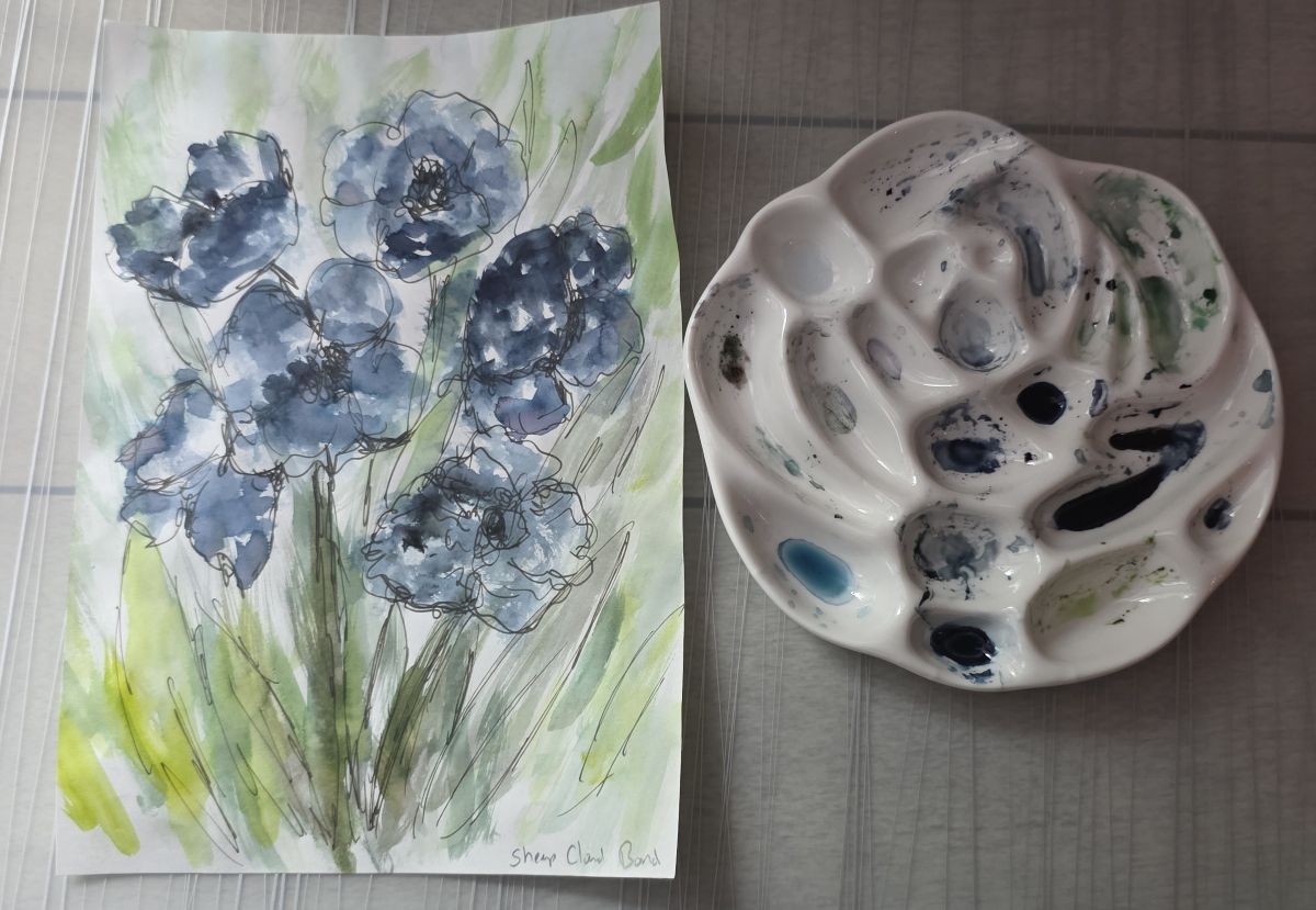 An ink painting of abstract blue flowers with green foliage and a ceramic paint palette with various colors of inks in the wells.