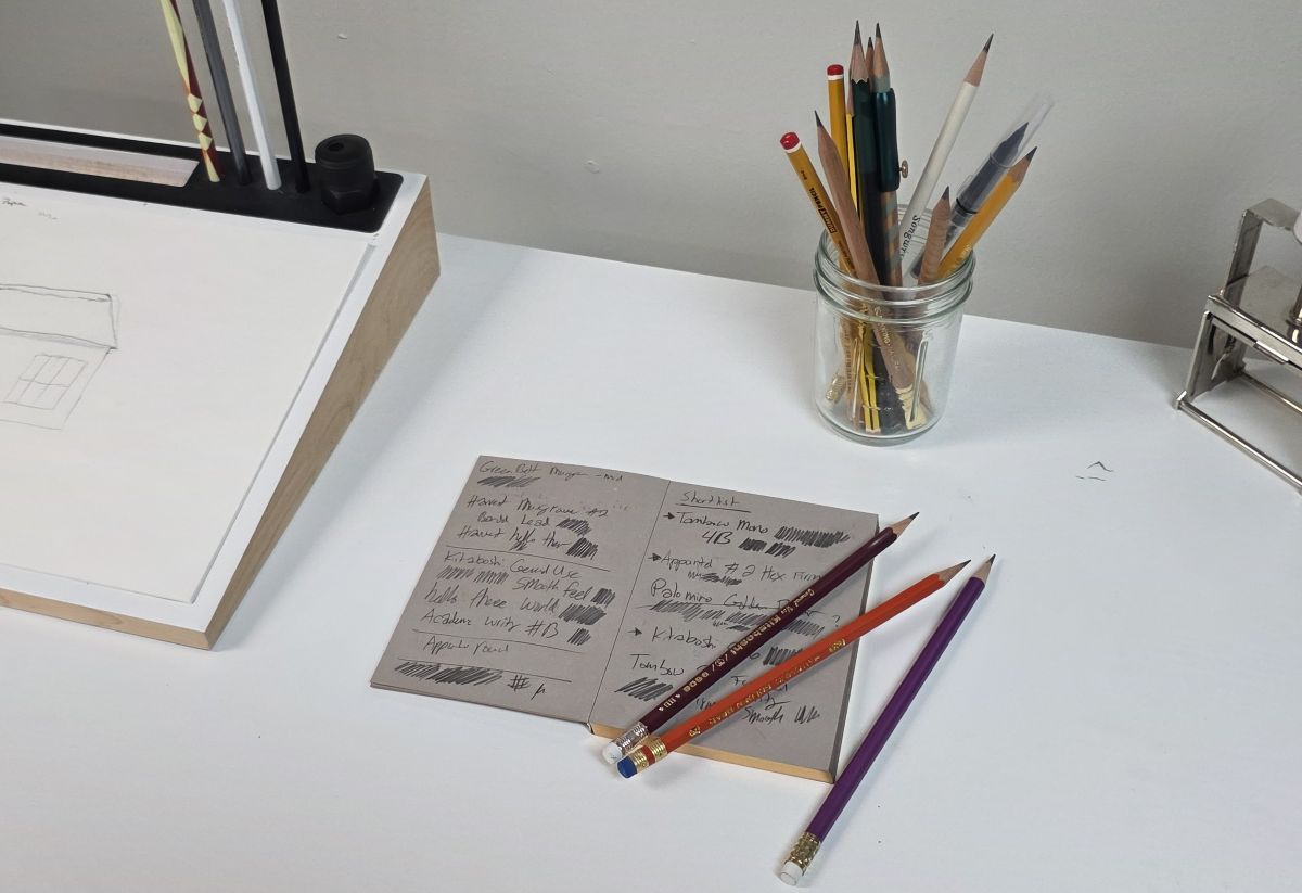 The testing desk at The Gentleman Stationer shop with a Blackwing pencil testing display, a glass jar full of sharpened pencils, a small notebook with medium gray paper opened to a page of pencil writing and three sharpened wood pencils on top.