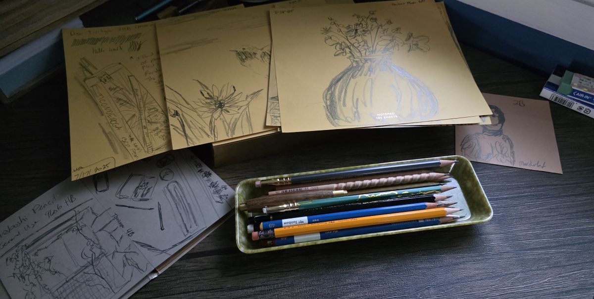 A shallow rectangle tray filled with sharpened wood pencils; several sheets of large, square, yellow paper with pencil sketches of a vase of flowers, a hummingbird and flower, and a box of pencils; a small notebook with medium gray paper open to pencil sketches looking out a window and items on a desk; and a small, square piece of blush paper with a pencil sketch of Murderbot.
