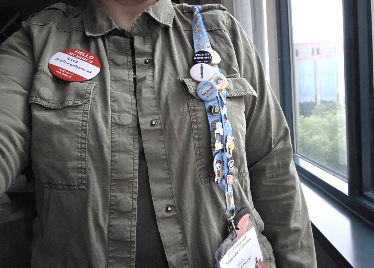 A person wearing an olive drab shirt-style jacket with a red and white button pinned on the left that says 'Hello My Name is Lisa, @oliveoctopus.ink and I am a Pen Addict!' and a lanyard full of other pins and buttons attached to a shoulder epaulet on the right with a single Rickshaw pen sleeve and name badge attached to the lanyard's clip.