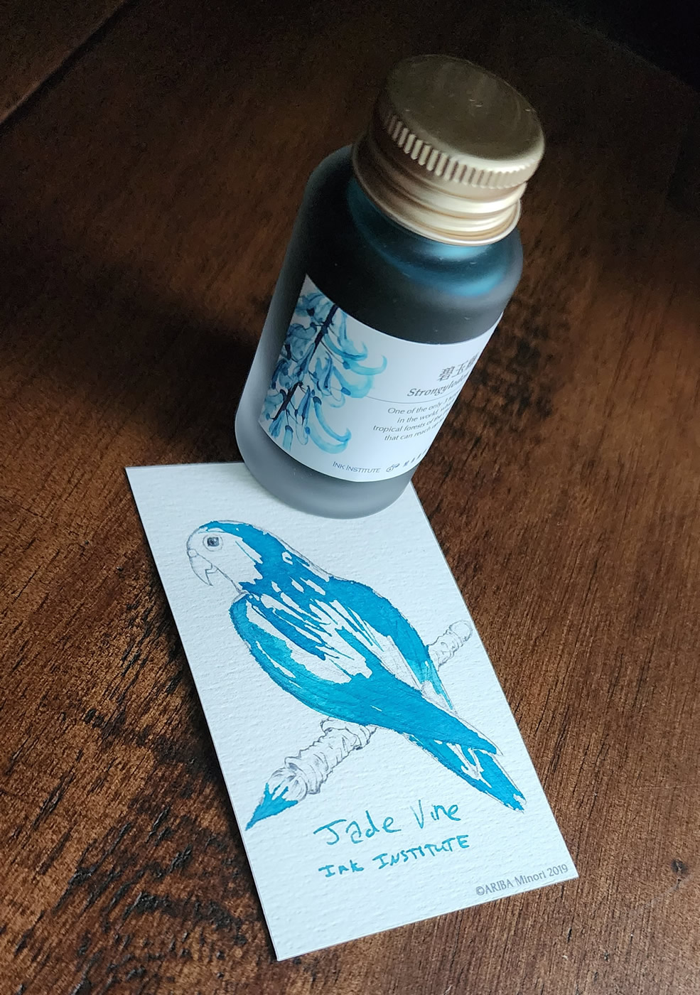A bottle of Jade Vine ink next to a small ink swatch card with a sketch of a bird holding a glass dip pen in its claws and bright teal ink highlighting the feathers.