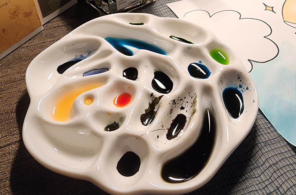 A ceramic paint palette in the rough shape of a flower with multiple sizes of paint wells filled with multiple colors of fountain pen inks being used for watercolor-style painting.