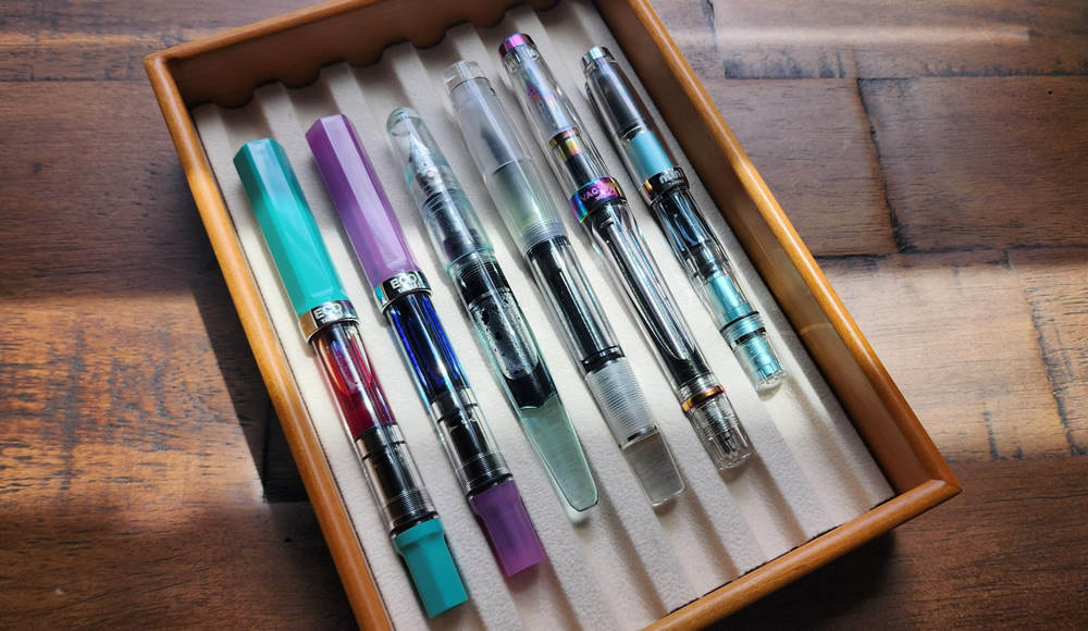 Six fountain pens that feature large open ink reservoirs in a lined wooden tray. Two TWSBI ECOs, a Franklin-Christoph P66 in Antique Glass, an Opus 88 Halo, a TWSBI Vac 700R Iris, a TWSBI Mini.