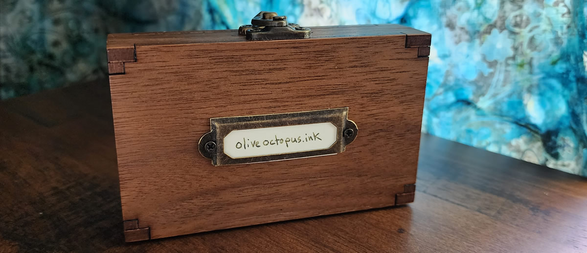 A small, thin wooden case with a metal clasp on top and metal nameplate on the front. 