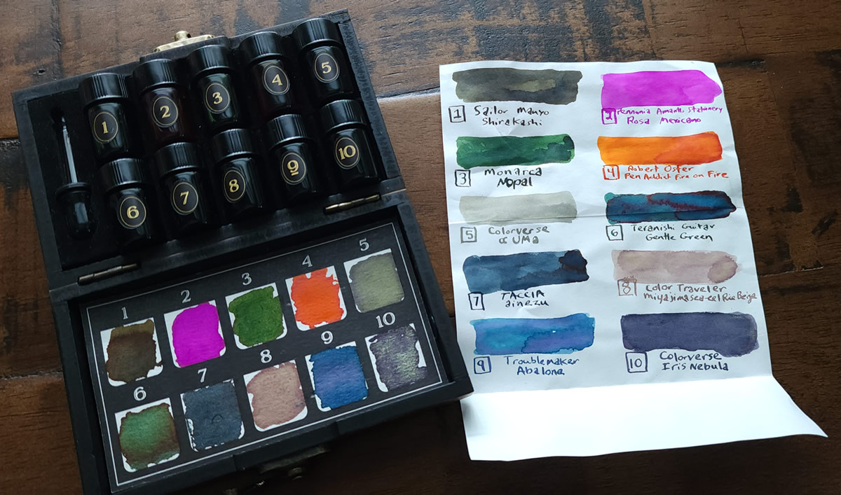 A small wooden case opened to show two rows of 5 small ink sample vials, with swatches of each color on a sheet in the other side of the case, next to a sheet of Tomoe River paper with each ink swatched and labeled.