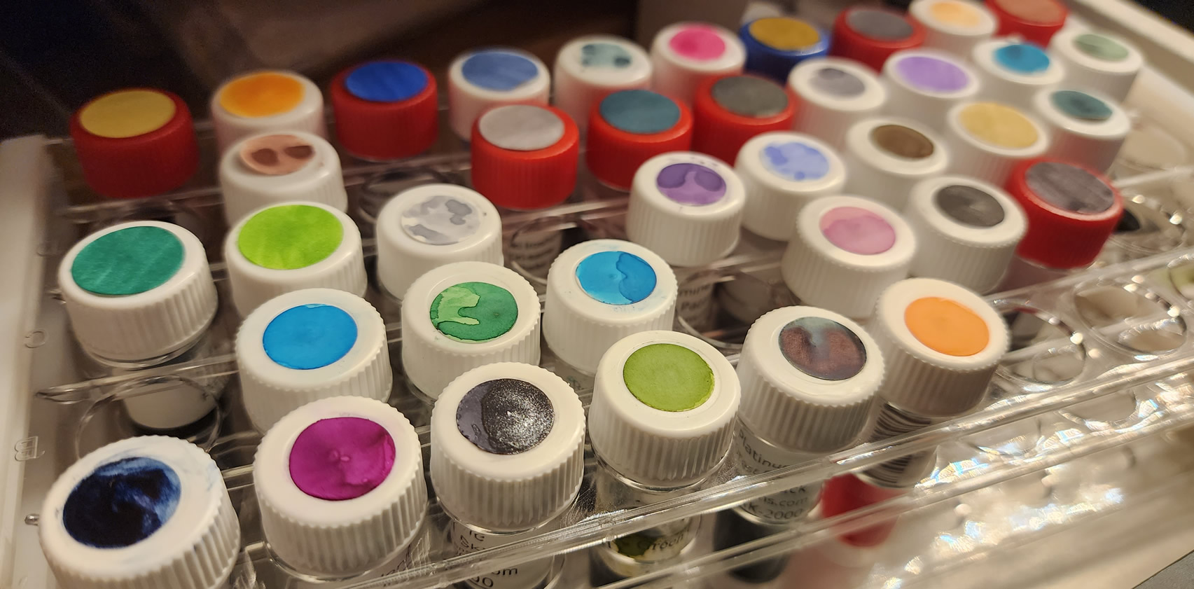 Several ink sample vials arranged in a plastic sample vial rack, each sample has a small round sticker with a swatch of the ink inside on top of the cap.
