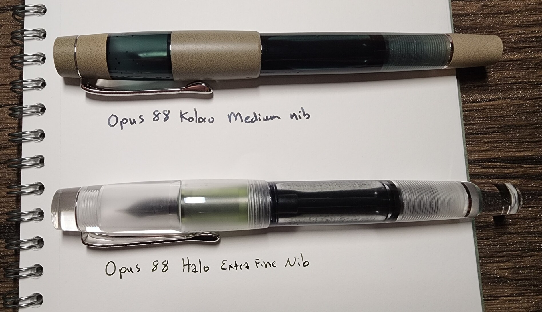 An Opus 88 Koloro pen in transparent teal acrylic/tan ebonite with a writing sample of the medium nib, and a transparent Opus 88 Halo with a green section next to a writing sample of the extra fine nib.