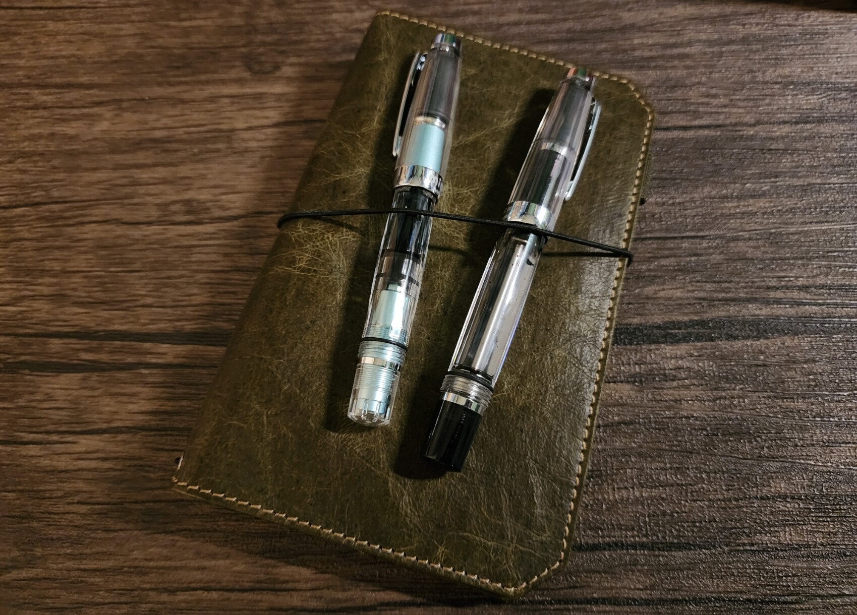 A TWSBI Diamond Mini AL (Mint Blue) and a TWSBI Mini Vac (Smoke) tucked into the elastic band around a passport-size Franklin-Christoph TN notebook cover in Olive NWF material.