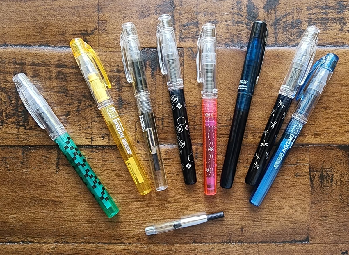 Eight different Preppy, Preppy Wa, and Prefounte transparent fountain pen models in green with black squares, yellow, clear, black with a geometric floral design, pink with sakura blossoms and wavy dotted lines, dark blue Prefounte model with a metal clip, blue-black with sparkly gold and silver dragonflies, and bright blue, plus a compatible silver-trim Platinum converter.
