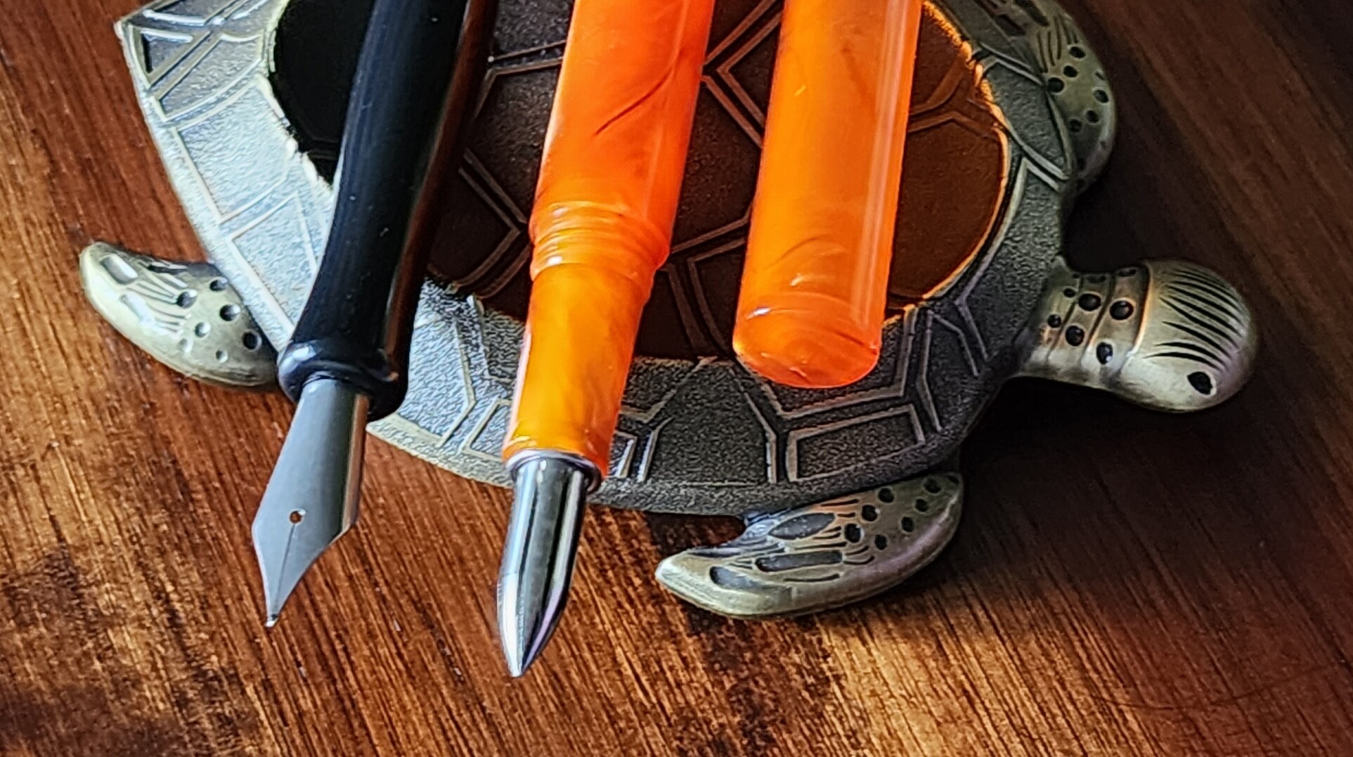 A Kakimori pen nib, shaped like a firm fountain pen nib without a feed, in a black plastic nib holder. A Kakimori stainless steel nib that is a conical shape with groves to hold ink in a ferrule that happens to fit ok into another plastic orange pen body with a cap. Both pens are resting on a metal turtle pen rest.