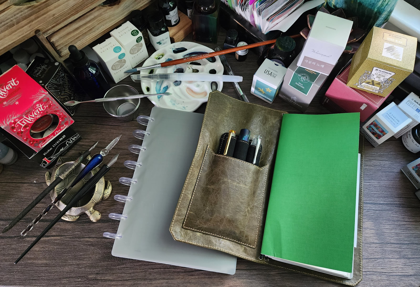 A top-down view of a desk with a Franklin-Christoph TN-size notebook cover with inserts, a 3 pen holder visible in the front cover pocket, on top of a gray TUL discbound Jr notebook with clear disks, surrounded by fountain pen ink boxes and bottles, several dip pens with metal and glass nibs, a paint brush, water brush and ceramic watercolor palette.