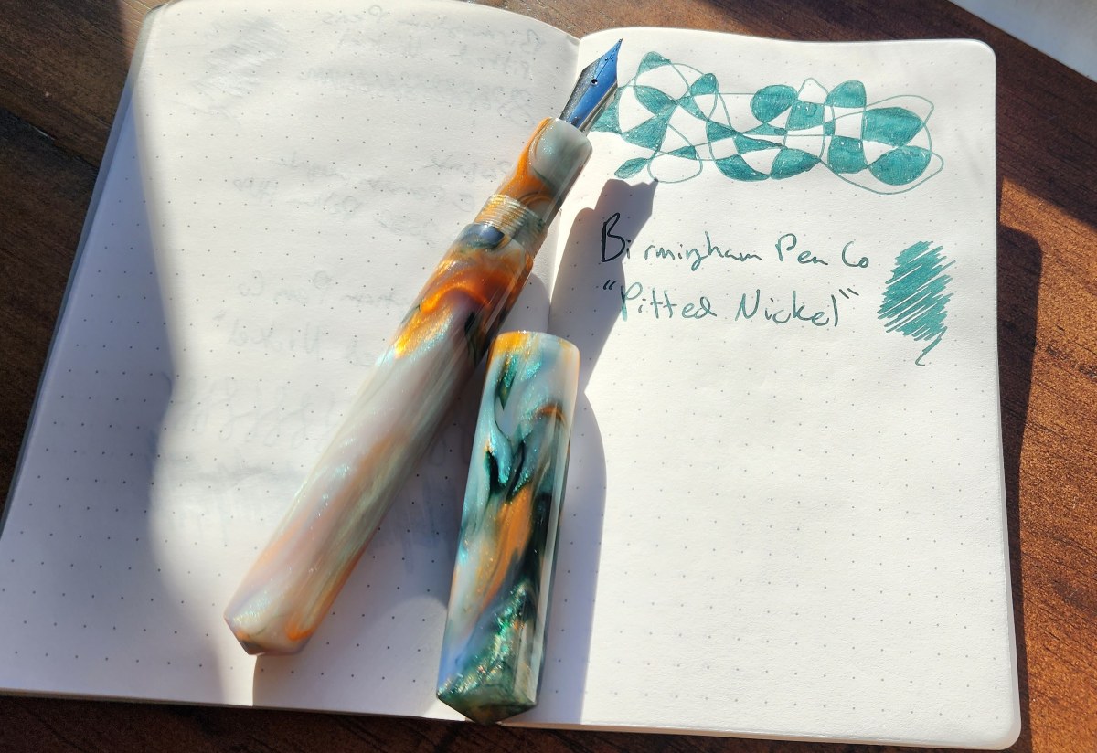 A notebook open to page with a doodle and written name for a patina green ink by Birmingham Pen Company called Pitted Nickel, with an uncapped fountain pen made from resin with pearlescent white, orange, and teal green swirls.
