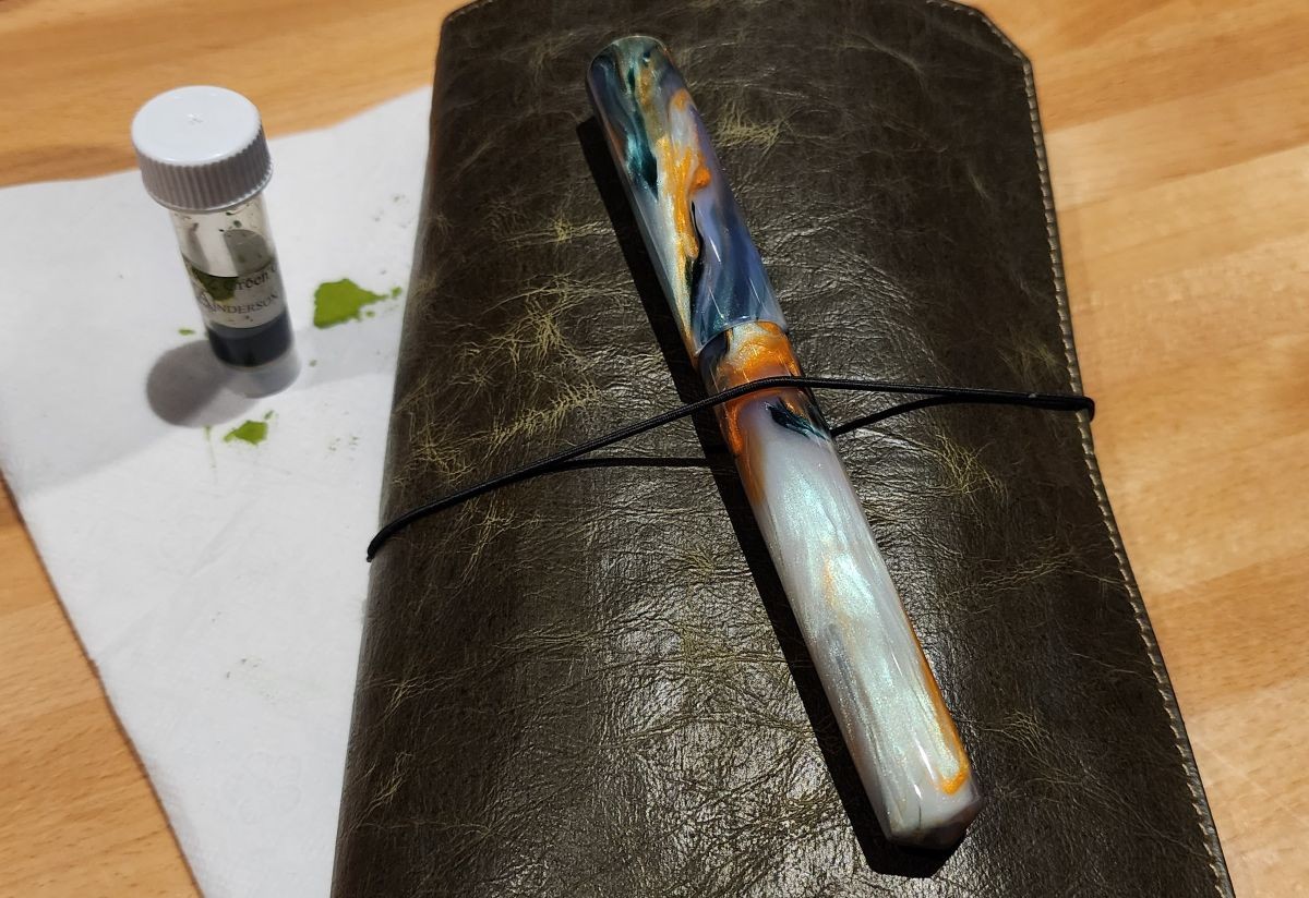 A Franklin-Christoph Vagabond A5-slim size notebook cover in dark green NWF material with a turned resin fountain pen on top, next to an ink sample vial with green ink.