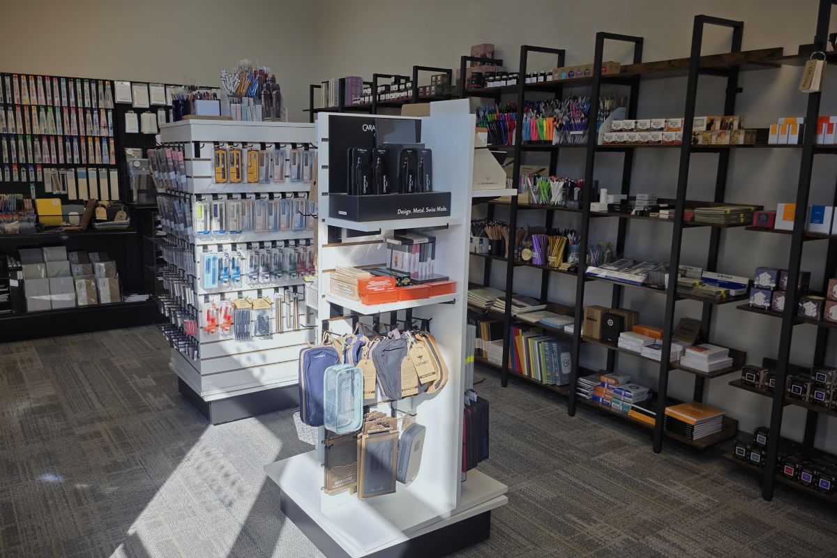 View inside The Gentleman Stationer shop with a spinner of Lamy pens and pencils, multiple shelves along the walls with pencils, paper, notebooks, planners, multipens, highlighters, etc. and two freestanding spinners with dip pens, stamps, fountain pens, gel pens, ballpoint pens, sharpeners, and other writing tools and stationery.