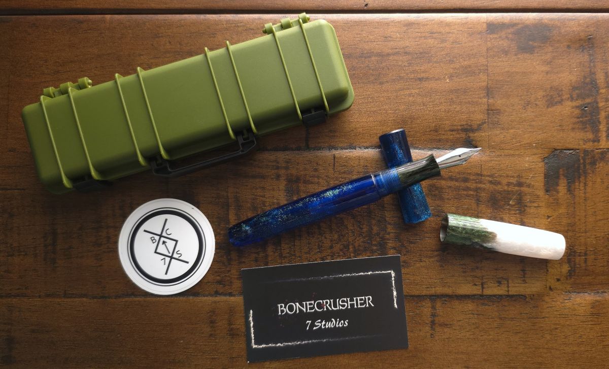 An uncapped fountain pen with a translucent blue body with green shimmer, a dark green section, and a cap that goes from dark green to bright white, resting on a pen rest next to a card and sticker for Bonecrusher 7 Studios and an army green plastic storage case.