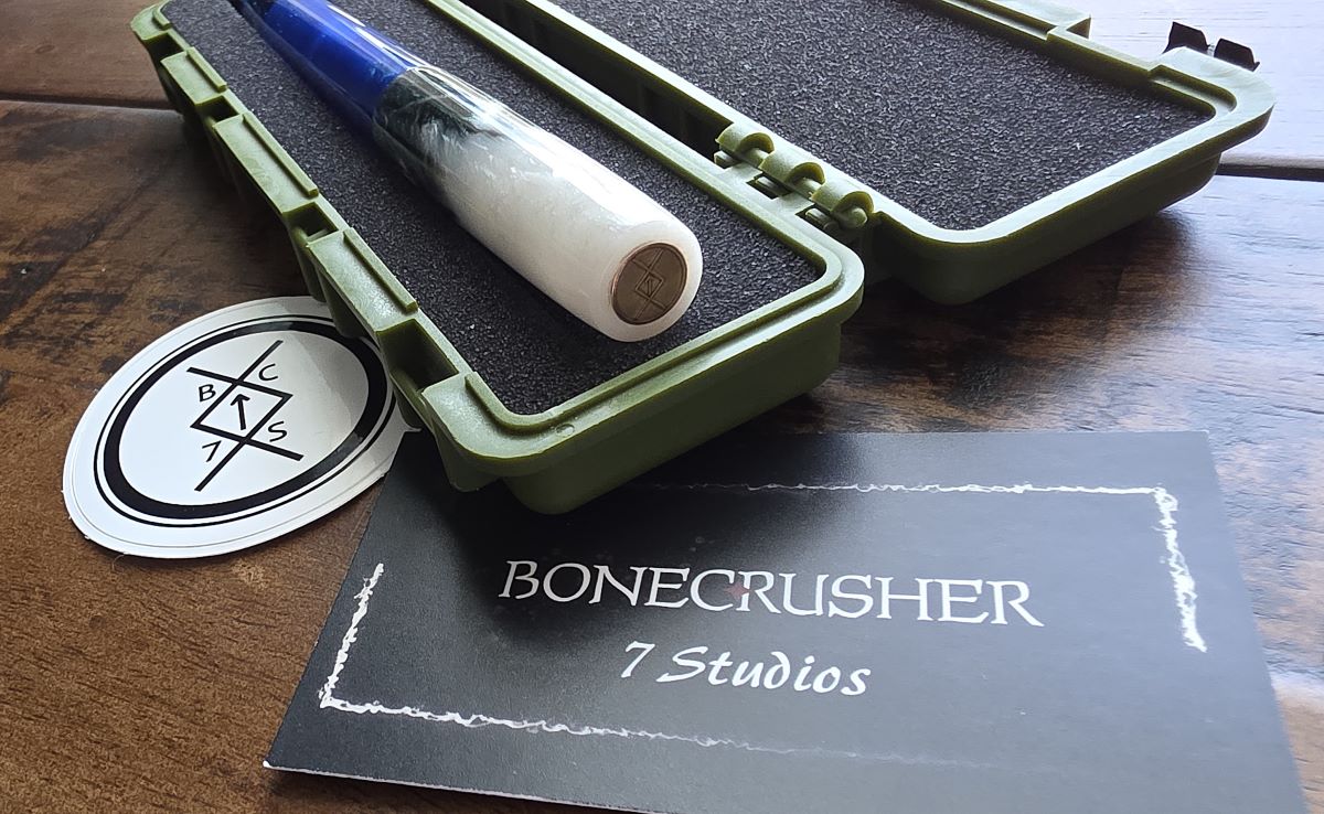 A closeup of the copper finial with the Bonecrusher 7 Studios logo on the top of the cap of a resin fountain pen with a green and white cap and translucent blue body, resting in an open plastic storage case lined with foam, next to a business card and sticker.