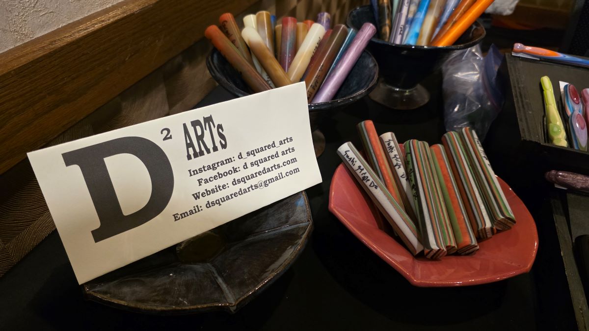 A business card for D² Arts with contact information, displayed in a handmade ceramic dish next to other ceramic dishes holding resin pen blanks.
