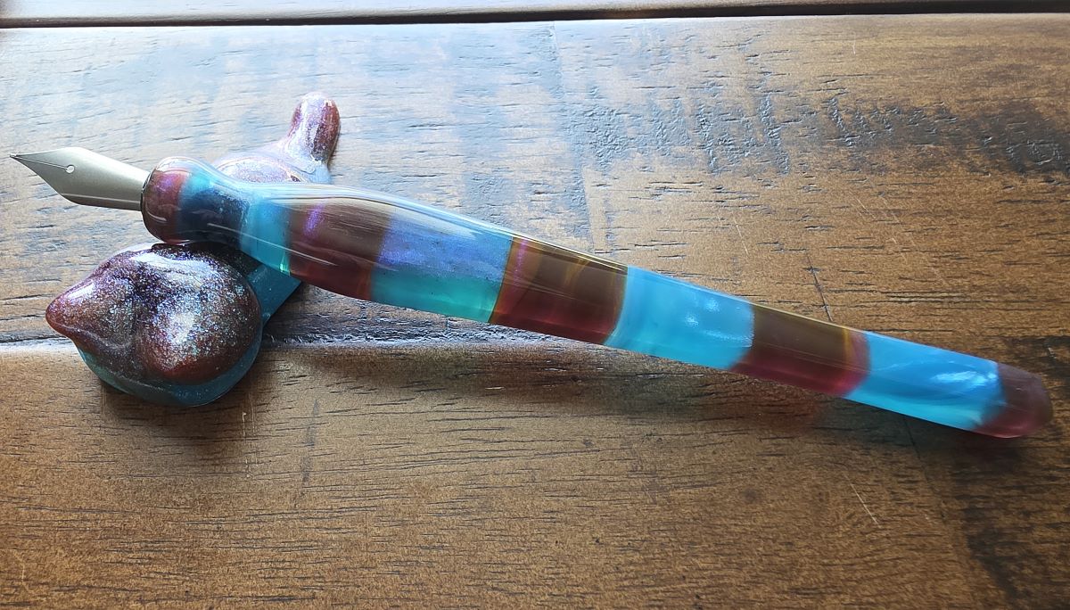 A dip nib holder of hand-carved resin with alternating wide sections of clear bright blue and warm brown resin with hints of purple shimmer, fitted with a Kakimori pen nib.