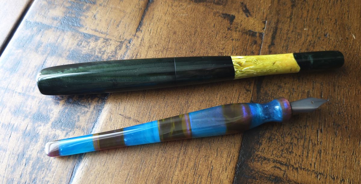 A long, dark green, hand-carved pen with a section of light green in the tapered body next to a hand-carved dip nib holder with metal nib with a pinched section and tapered squared body with colorful resin in alternating sections of dark brown and bright blue.