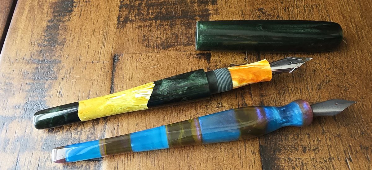 An uncapped dark green, hand-carved pen with a section of light green in the tapered body and a bright orange section next to a hand-carved dip nib holder with metal nib with a pinched section and tapered squared body with colorful resin in alternating sections of dark brown and bright blue.