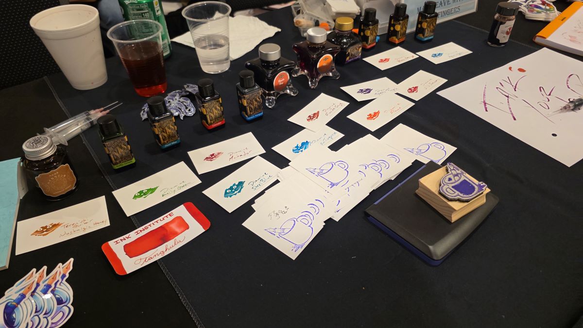 A line of ink bottles, swatches, swatch cards, a stamp, stickers, etc. at the ink filling table.