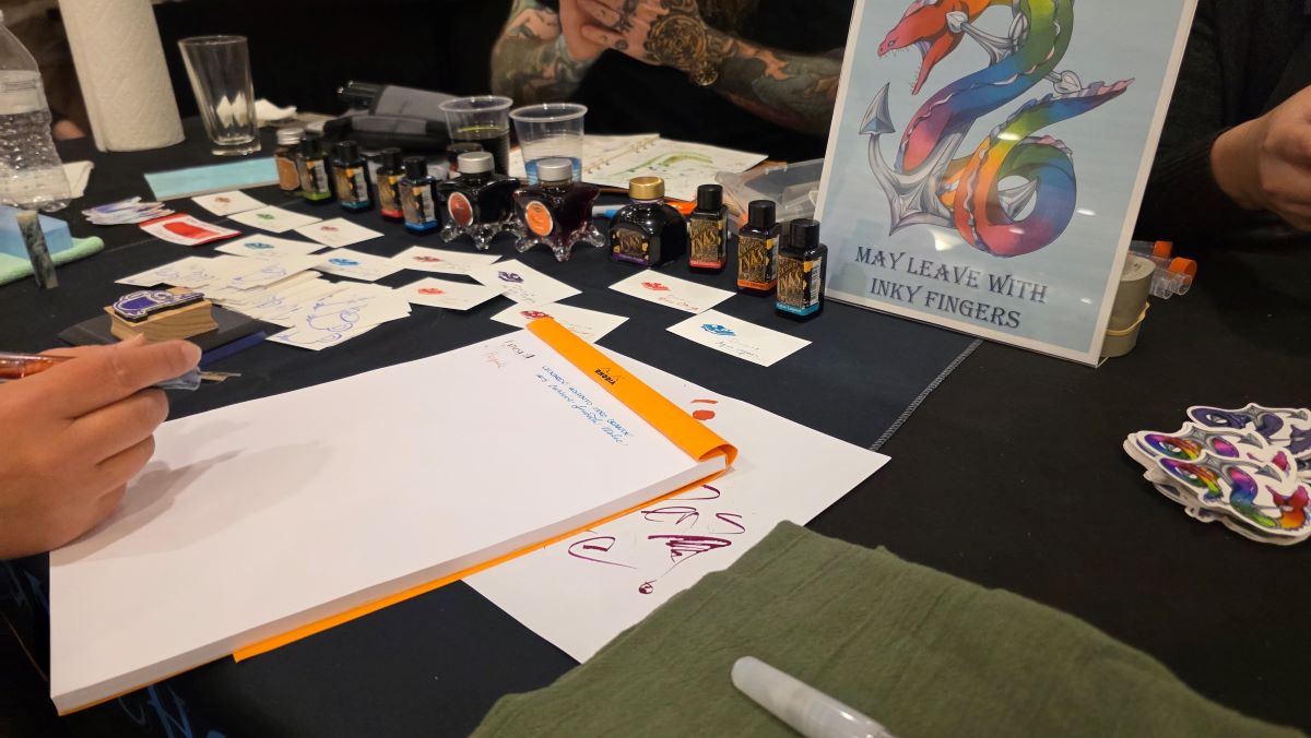 Someone testing out a newly inked pen on a Rhodia notebook, with bottles of ink in the background and a sign with the Eel and Anchor rainbow logo that says 'May Leave With Inky Fingers.'