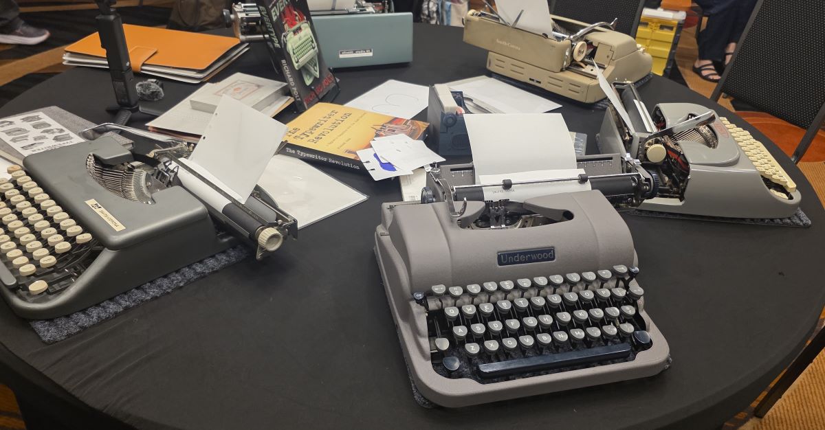 A large round table covered with 5 typewriters and related papers and books.