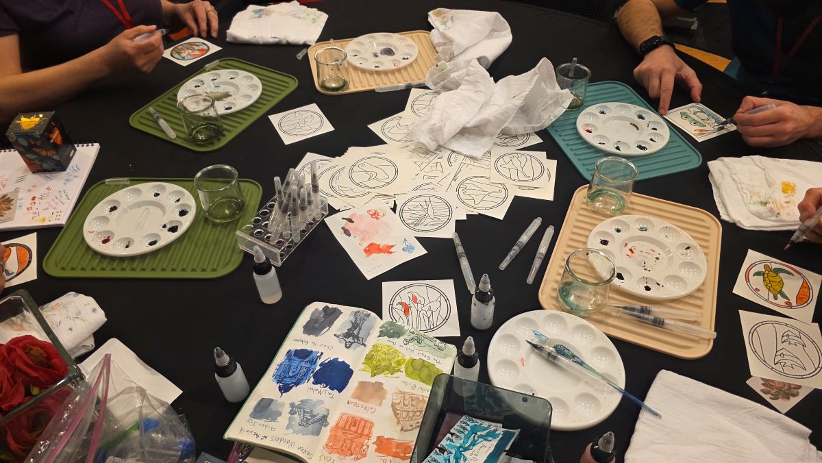 A large round table covered in ink palettes, coloring sheets, water brushes, towels, and people painting with inks.
