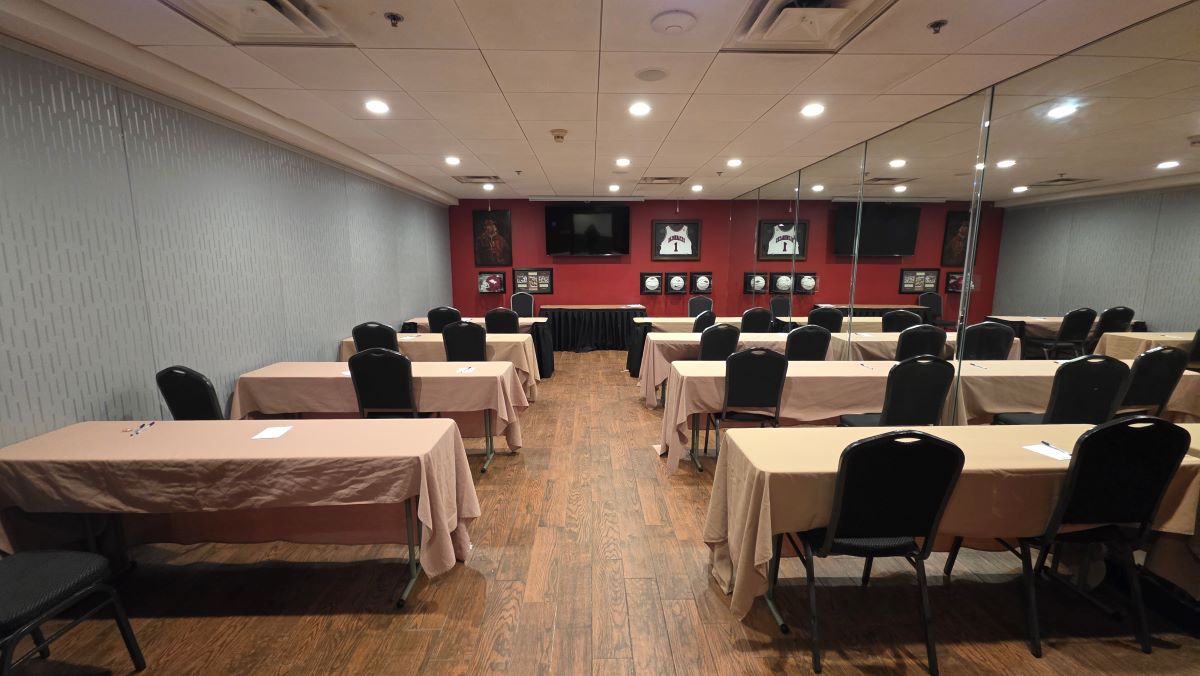 A small hotel meeting room with 8 narrow tables with two rows each, the back wall is red with Razorback memorabilia, and a wall on the right of all mirrors.