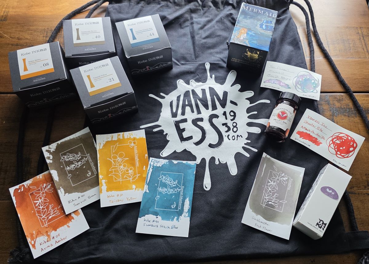 A selection of fountain pen inks and swatches on top of a black vanness1938.com drawstring bag including four Kobe inks, Van Dieman's Mermaid, Vanness Skeeter Bite, and Troublemaker Frost Flower.