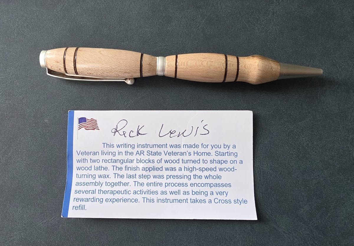 A hand-turned pen with a curved design in light wood with dark accent bands and light silver hardware, and a card explaining how it was made by a Veteran living in the AR State Veteran's Home.