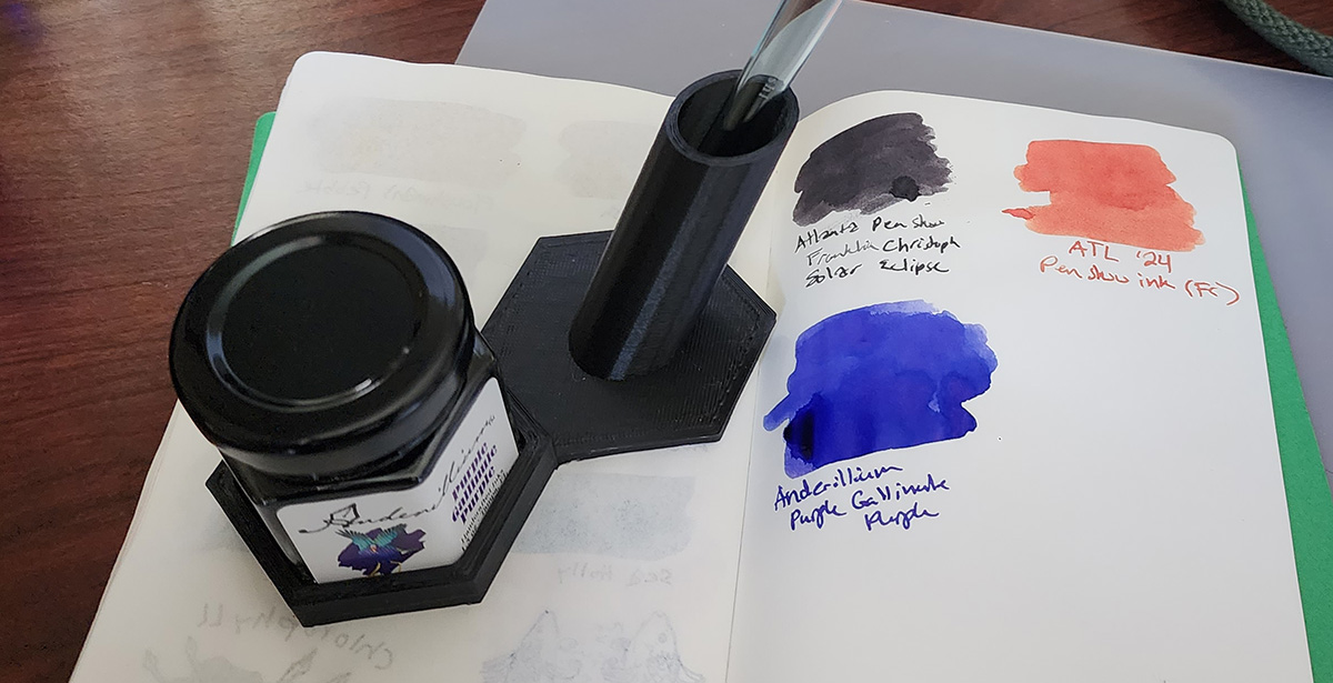 Notebook showing swatch of the bright blurple Anderillium Purple Gallinule Purple ink, and a 3D printed custom stand holding the hexagon-shaped bottle and a dip pen.