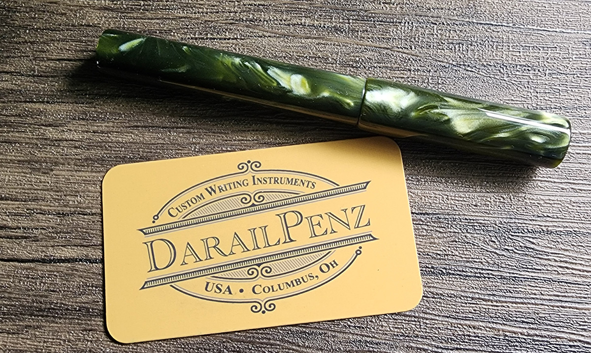 Chatoyant green resin fountain pen with straight sides and flat ends, next to a card for Derail Penz Custom Writing Instruments, Columbus, Ohio.