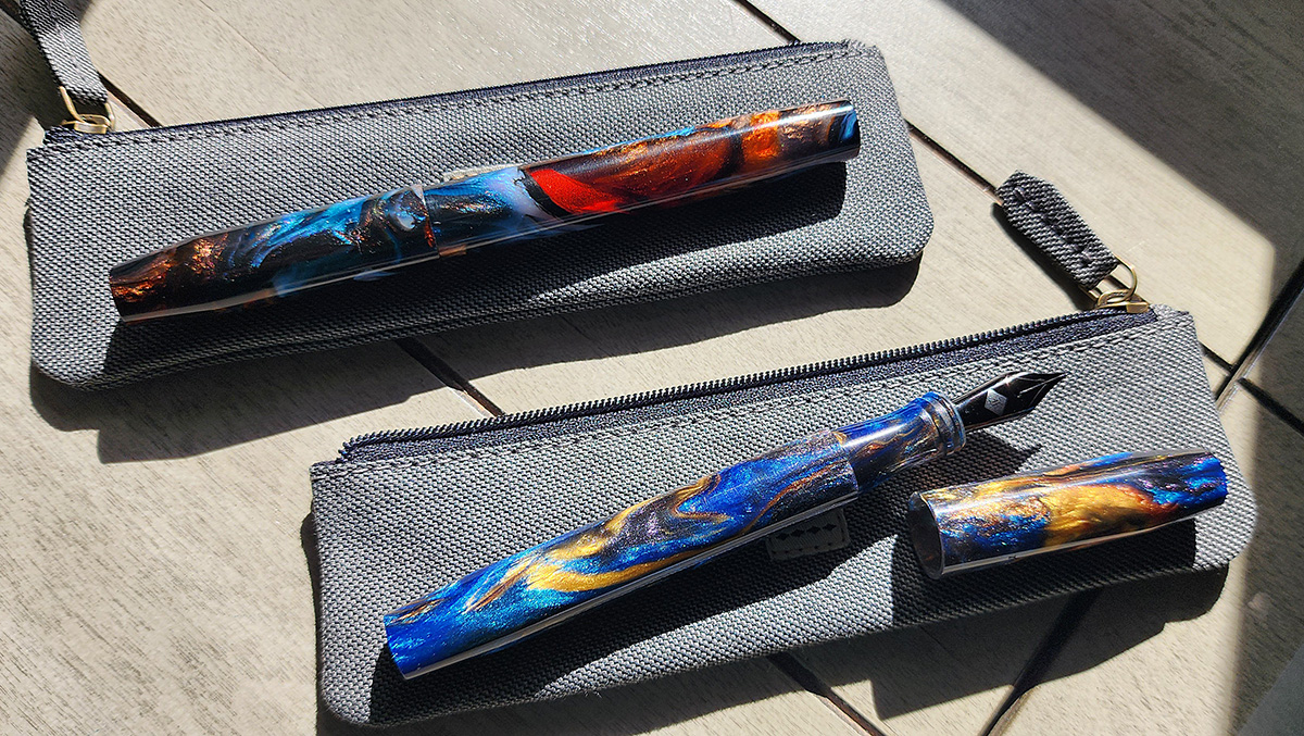 Two Franklin-Christoph Model 46 pens resting on gray zipper pouches, one pen with swirling transparent orange and frosty blue, and the other with golden yellow swirls in a sea of dark blue and purple shimmer.
