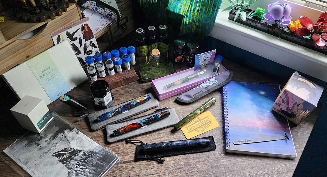 Multiple pens, ink bottles, ink samples, notebooks, and accessories laid out across the desk.