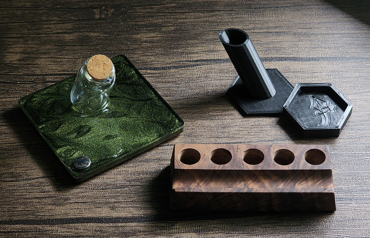 Handmade pen holder that is a green resin square with angled glass bottle to hold a pen, a wooden stand with spots for 5 sample vials and a horizontal pen rest, and a 3D printed stand for the hexagon Anderillium bottle and a pen.