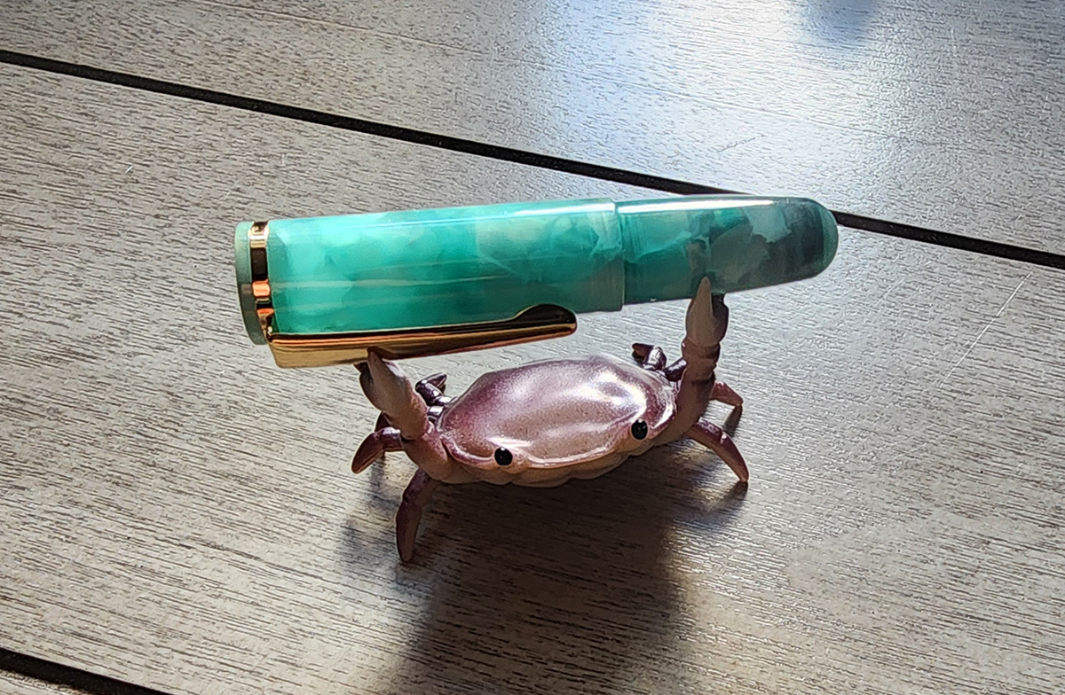 A crab pen holder with a small 2.5 inch long mini pen in a bright turquoise green material.