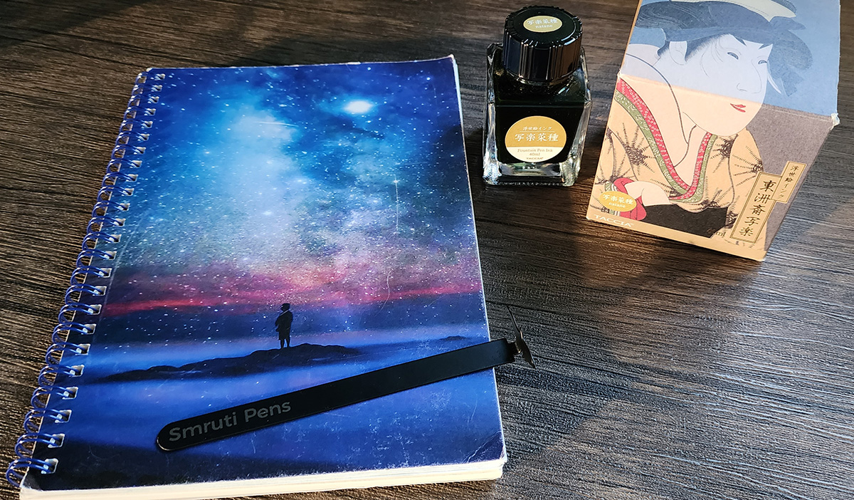 A ring-bound A5 notebook with starry night sky cover and bottle of golden yellow Taccia Natane ink.