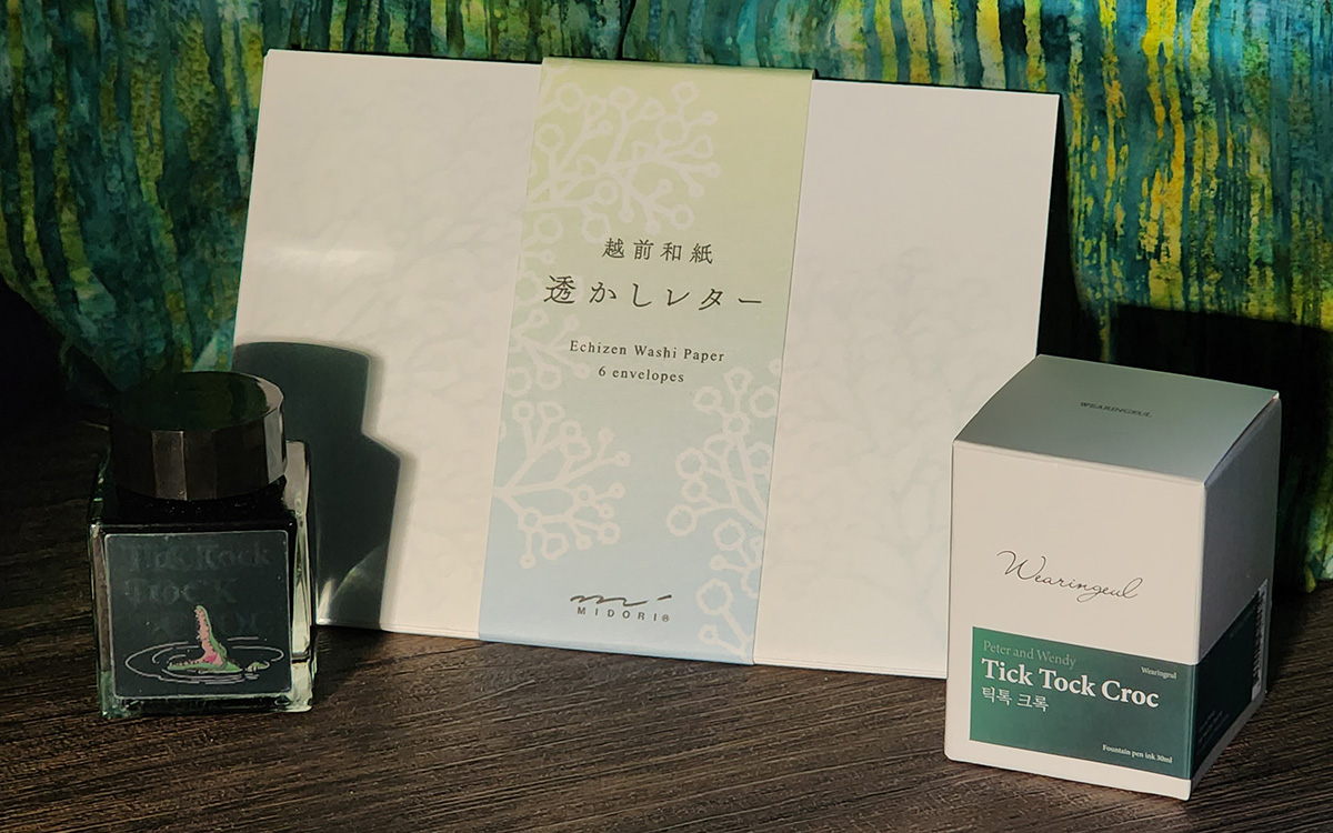 A pack of white Midori envelopes with watermark floral pattern and bottle of Wearingeul Tick Tock Croc ink, a blue-ish green ink in a square bottle with a lenticular label that shows the animated crocodile with wide open mouth.
