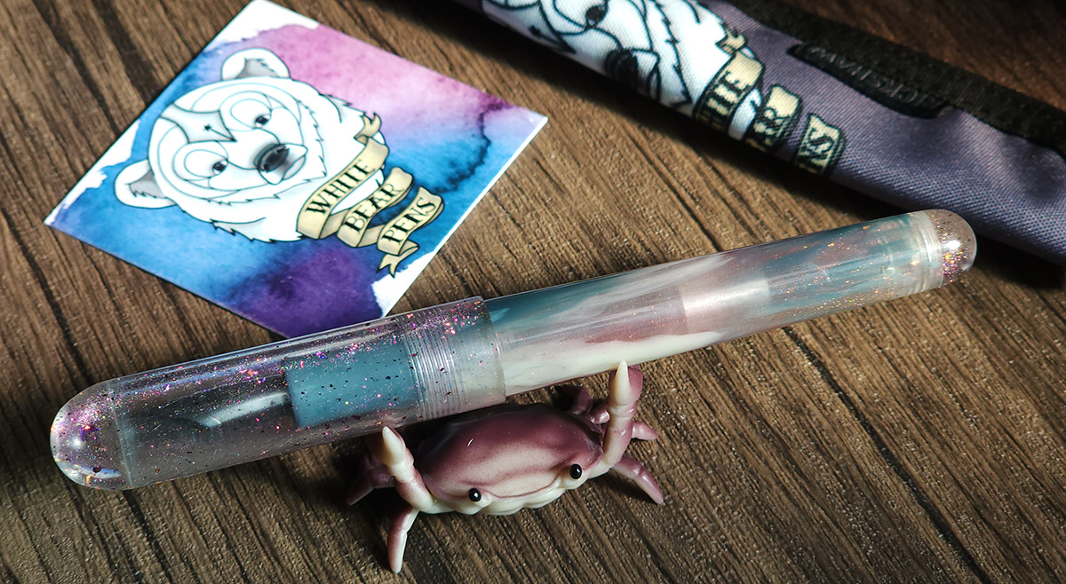 A resin fountain pen with clear domed cap sparkling with purple/pink/orange glitter and swirled blue gray, white, and translucent mauve body. White bear logo card and custom Rickshaw pen sleeve in background.