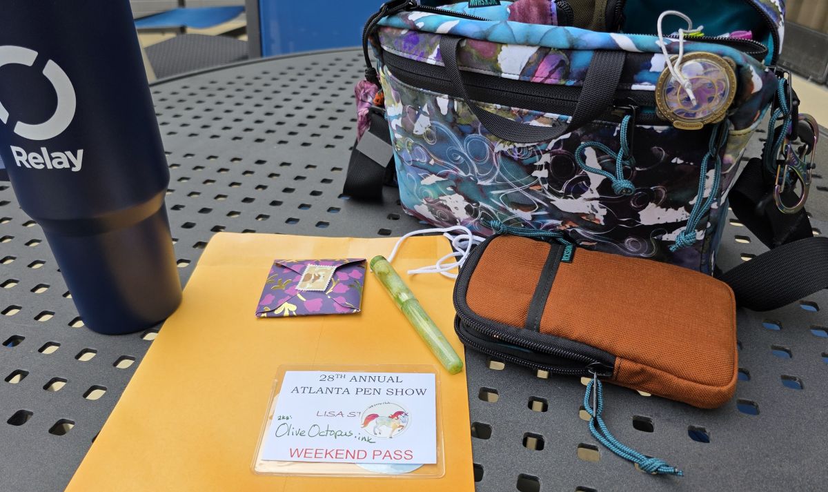 An outdoor table with an oversize mug with the Relay logo, a manila envelope, small square purple envelope, Atlanta Pen Show badge for Lisa, aka OliveOctopus.Ink, green Newton fountain pen, orange Rickshaw Sinclair pen case, Rickshaw Inktopus Banzai bag.