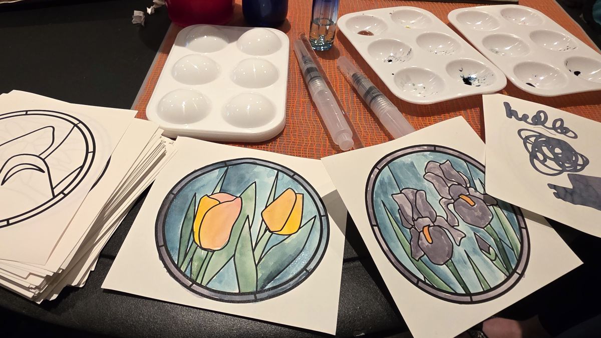 Plastic paint palettes with ink, water brushes, dropper bottles of water, a stack of coloring sheets, and some painted stained glass flowers.