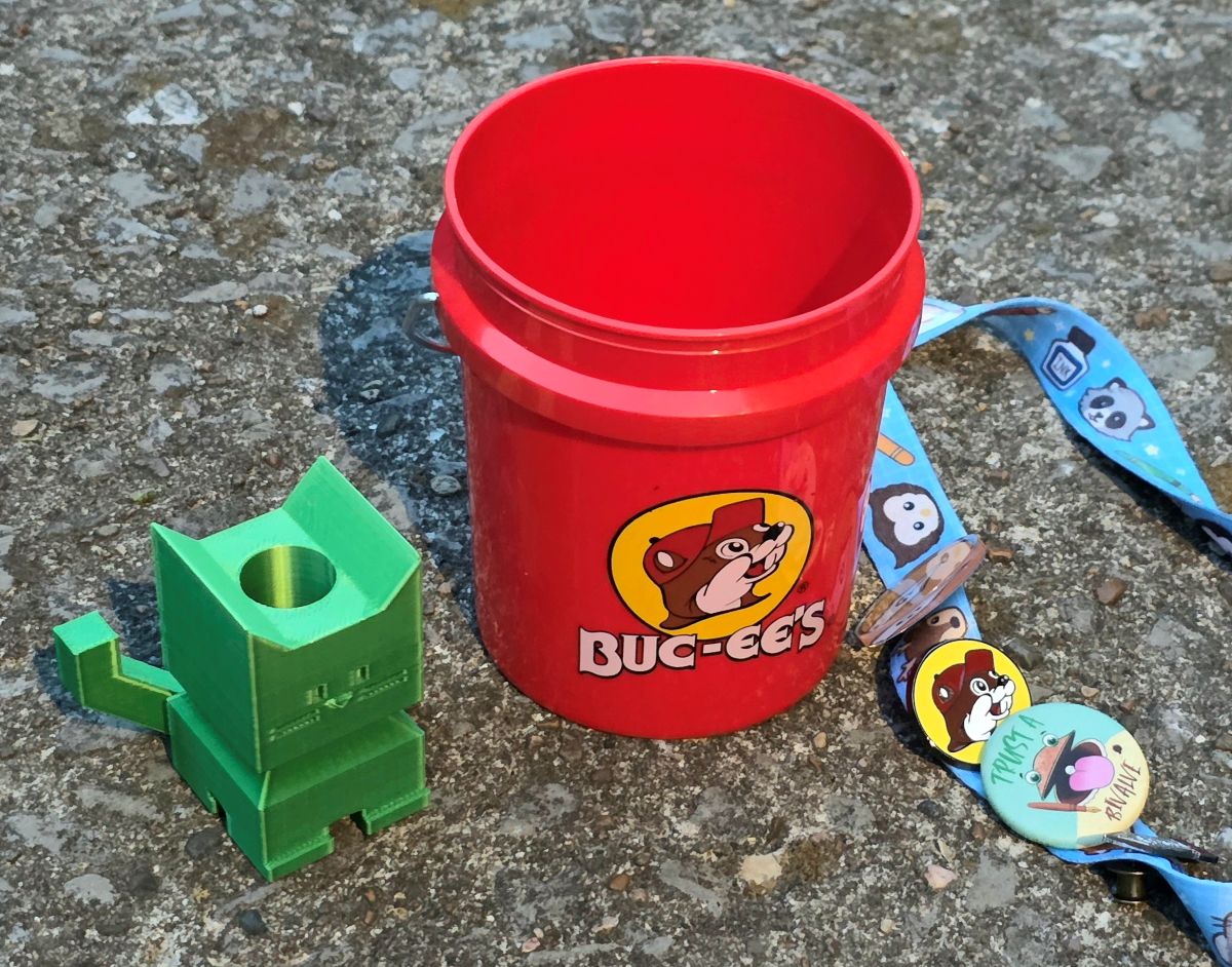 Small red Buc-ee's bucket, Buc-ee's pin on a lanyard with other pins, and a green cat-shaped ink vial holder.