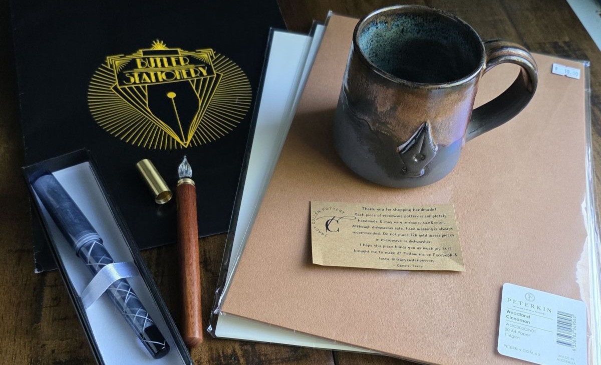 A stack of paper, handmade mug featuring a nib design, glass dip pen with wood handle and metal cap, and translucent gray fountain pen.