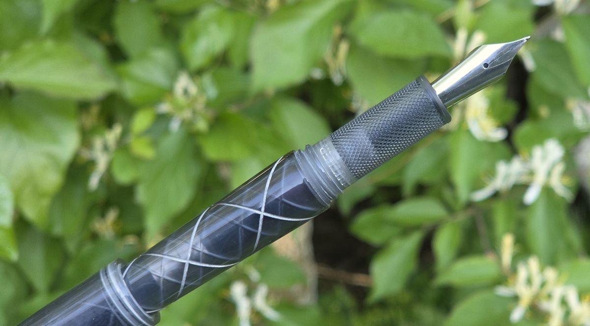 An uncapped fountain pen in a translucent gray with a knurled grip, and silver accent lines criss-crossing down the body.