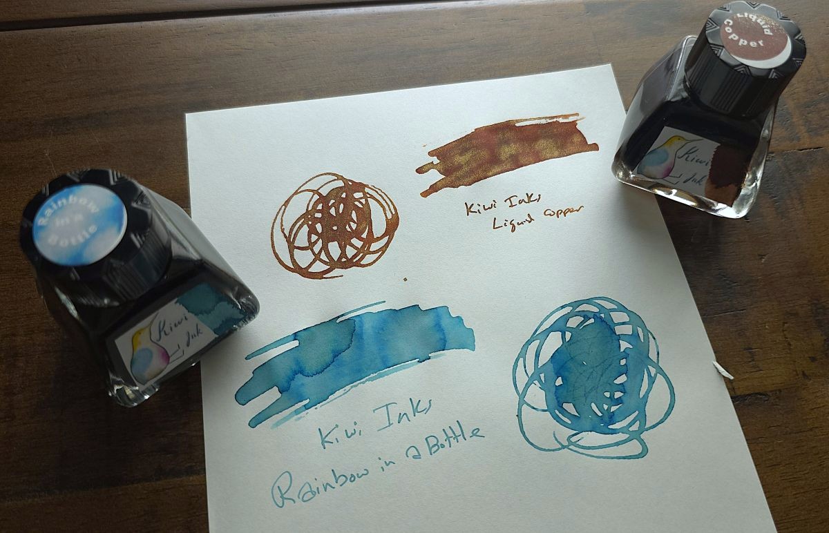 Glass fountain pen ink bottles and swatches for Kiwi Inks Liquid Copper, a medium brown with shimmer, and Rainbow in a Bottle, a soft chromashading blue.