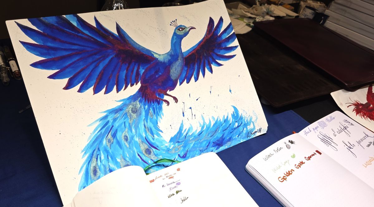 An ink painting of a peacock with spread wings with rich blue ink, red sheen highlights, and green shimmer feather details displayed on the Kiwi Inks table.