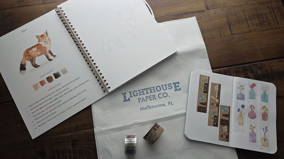 A watercolor workbook open to a page with a guide for painting a red fox on the left page and a blank page with the outline of the fox on the right, two rolls of washi tape including one with a travel/books theme with gold foil accents and another with colorful vases of flowers on a transparent background.
